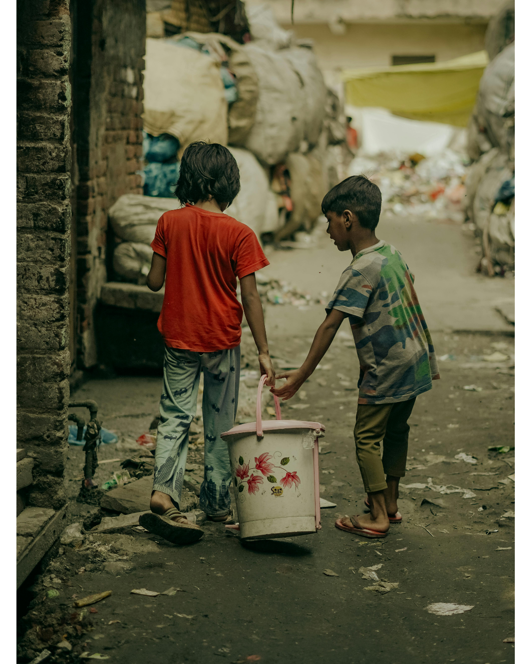 Two siblings walking together in a slum lane, one holding a small bucket — the kind of childhood Mayaar's orphanage appeal seeks to change